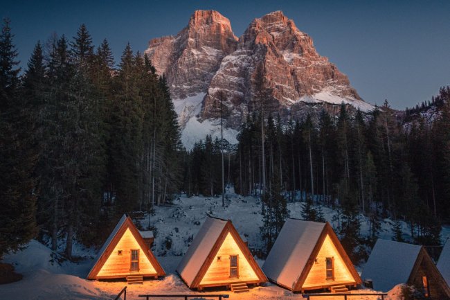 Baite illuminate nella neve, ai piedi di una montagna rocciosa al tramonto.