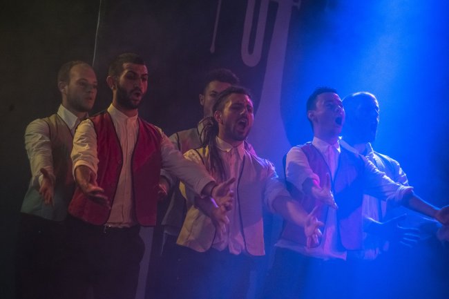 Gruppo di cantanti maschili sul palco con luci blu, vestiti formali.