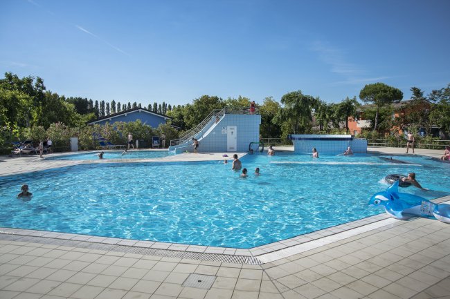 Piscina all'aperto con persone che nuotano. Sullo sfondo, un piccolo scivolo e alberi.