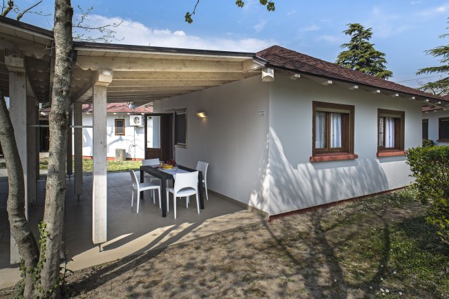 Casa con patio coperto, tavolo e sedie da esterno. Alberi e cielo azzurro sullo sfondo.