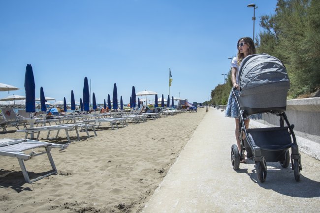 Donna con passeggino su una passeggiata vicino alla spiaggia, ombrelloni blu e sdraio sulla sabbia.