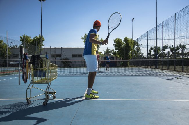 Giocatore di tennis colpisce la palla sul campo. Un carrello con racchette è vicino.