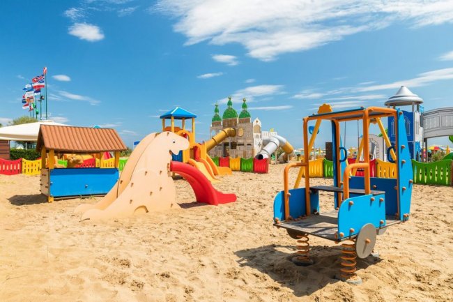 Area giochi sulla sabbia con scivoli colorati e strutture per bambini sotto un cielo azzurro.