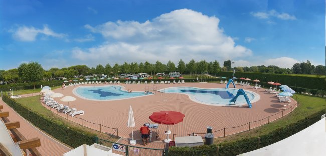 Piscina all'aperto con persone, ombrelloni e alberi sullo sfondo.