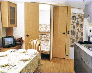 Cucina con tavolo apparecchiato, televisione e porta aperta verso altra stanza.